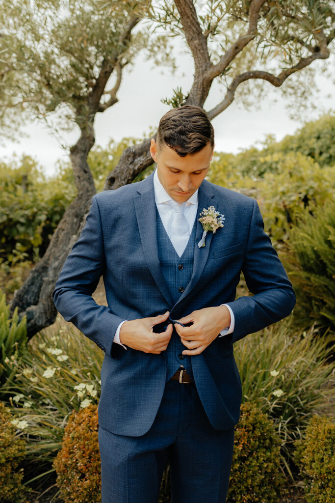 A man in a blue suit adjusts his jacket outdoors, standing in front of greenery and a tree, with a serious expression.