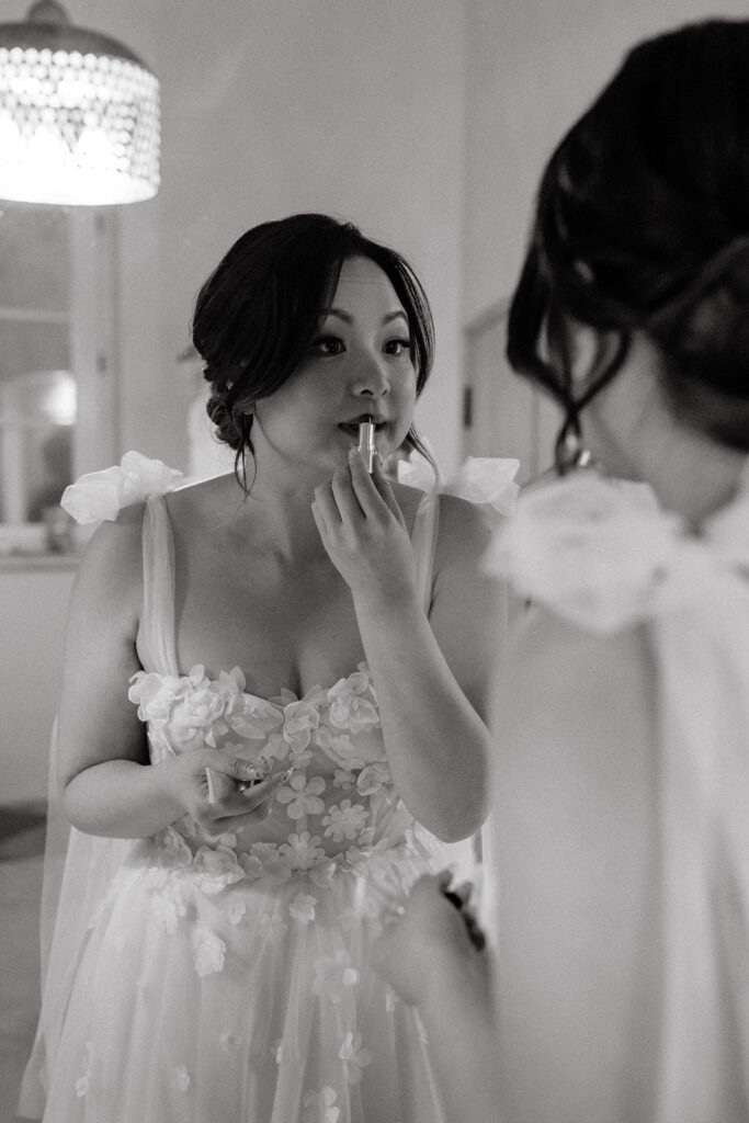 A woman in a floral dress applies lipstick while looking in a mirror.