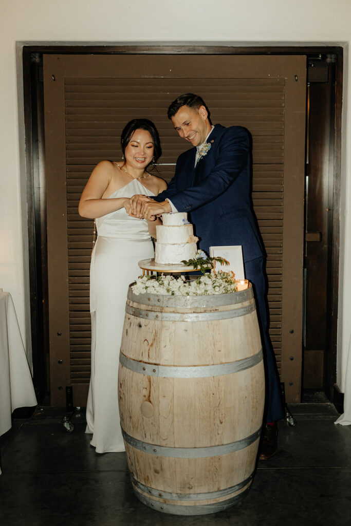 A couple in formal attire cuts a small tiered wedding cake placed on a wooden barrel.
