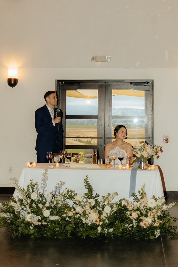 A man in a suit gives a speech with a microphone beside a seated woman at a floral-decorated table. Candles and drinks are on the table, with a door and scenic view in the background.