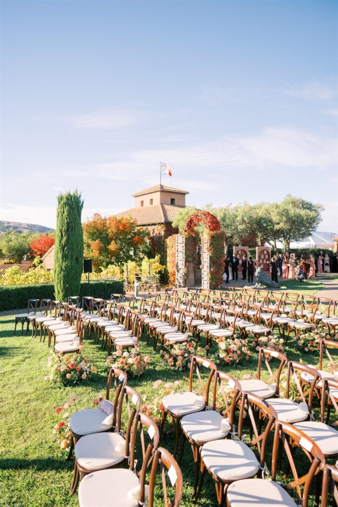 Outdoor wedding setup with rows of wooden chairs and floral arrangements on a lawn. A building with a flag and guests in formal attire are visible in the background.