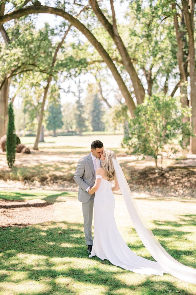 A bride and groom stand together on a sunlit, grassy area surrounded by trees. The bride wears a long white dress with a veil, and the groom is in a light gray suit.