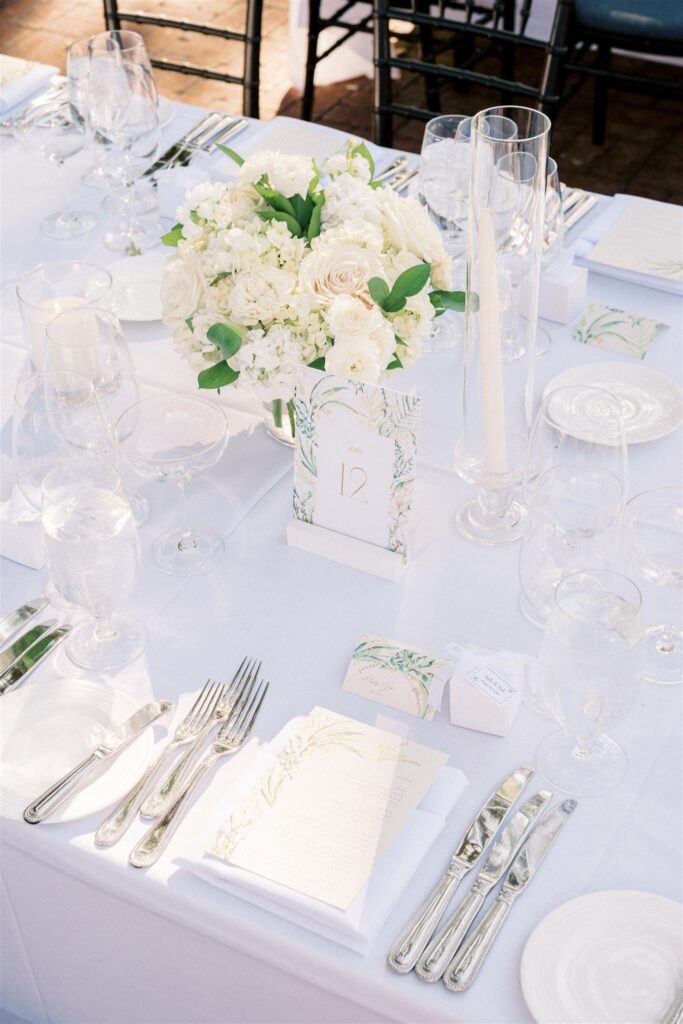 Elegant table set for a formal event with white flowers, candles, glassware, silver cutlery, and a table number 12. A white menu with floral design is placed near the center.