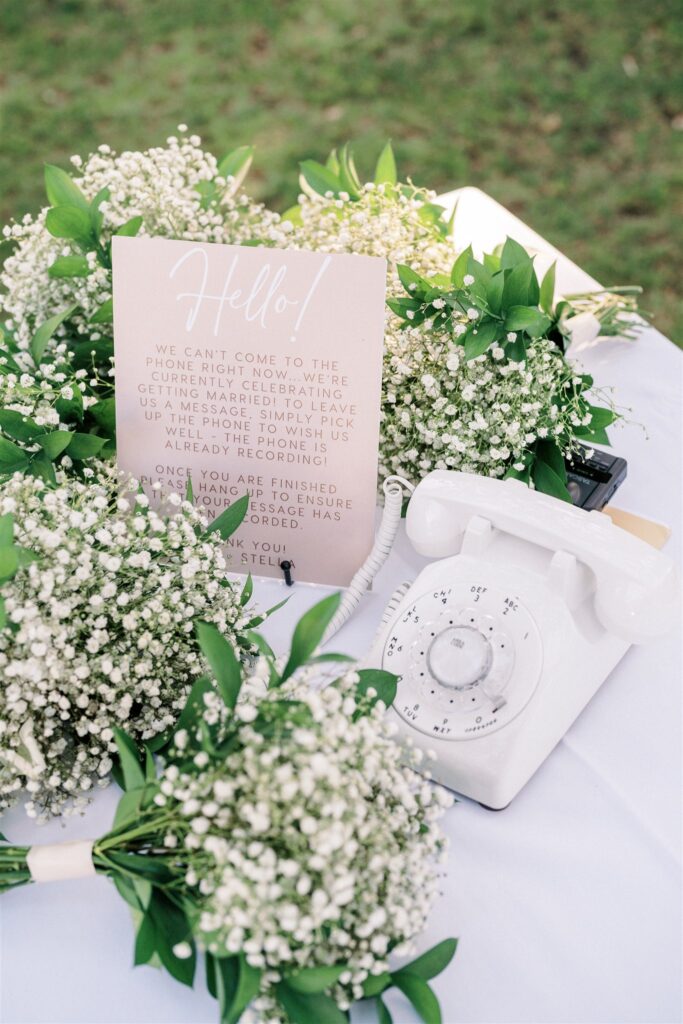 A white rotary phone sits on a table surrounded by white flowers and greenery. A sign with instructions for guests rests among the flowers.