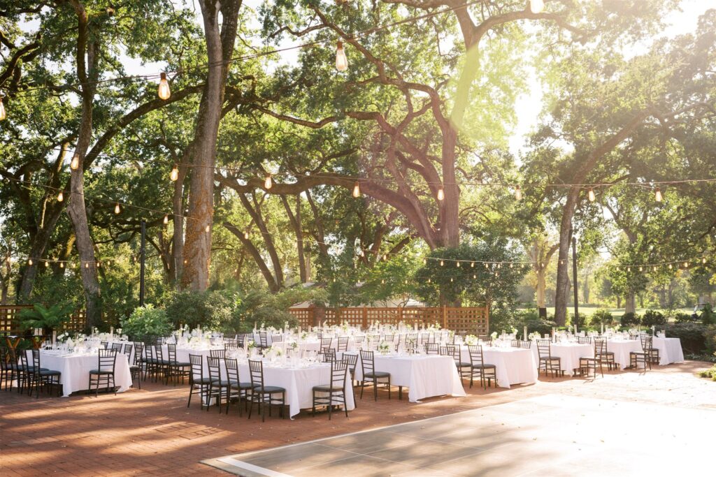 Outdoor event space with tables and chairs set under large trees. White tablecloths and string lights create a festive atmosphere. Sunlight filters through the branches.
