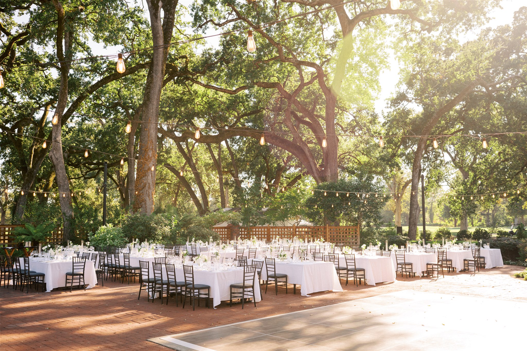 Outdoor event space with tables and chairs set under large trees. White tablecloths and string lights create a festive atmosphere. Sunlight filters through the branches.