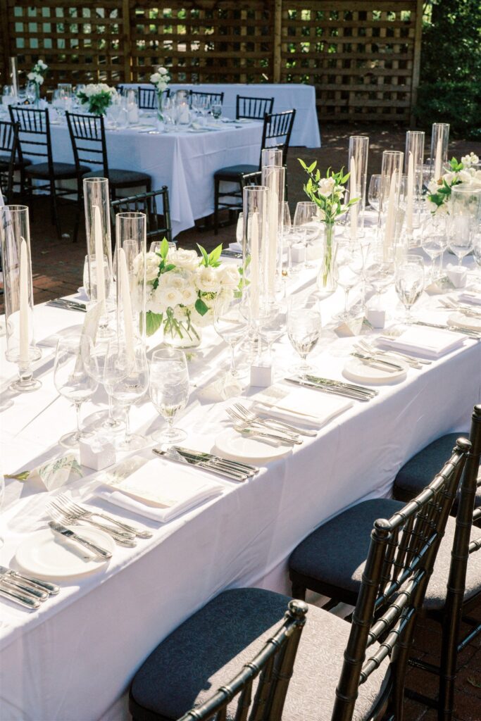 Long dining tables with white tablecloths are set with glassware, tall candles, and flower arrangements. Black chairs surround the tables, and sunlight casts shadows across the setup.