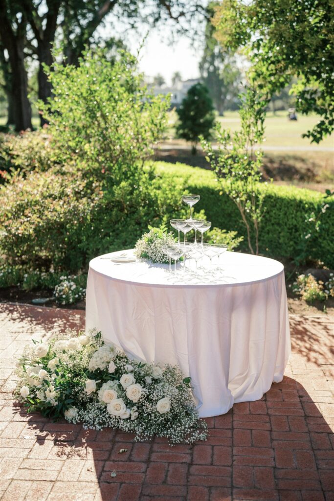 A round table with a white tablecloth holds a champagne tower. White roses and greenery are arranged at the base. The setting is outdoors with greenery in the background.