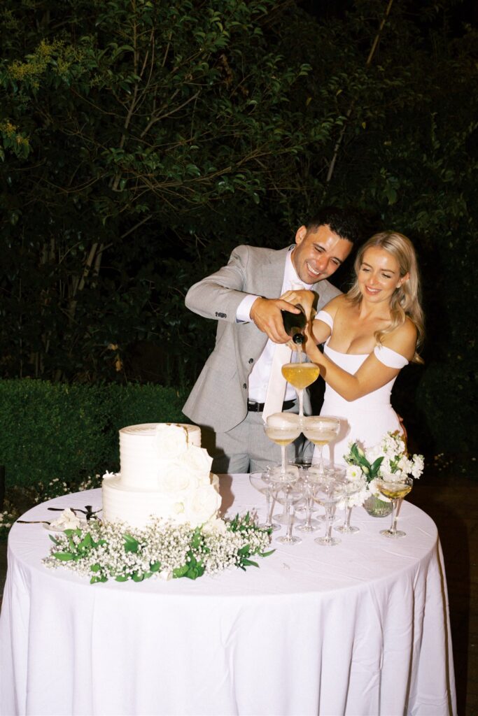 A couple in formal attire pours champagne into a glass tower next to a white cake on a round table outdoors.