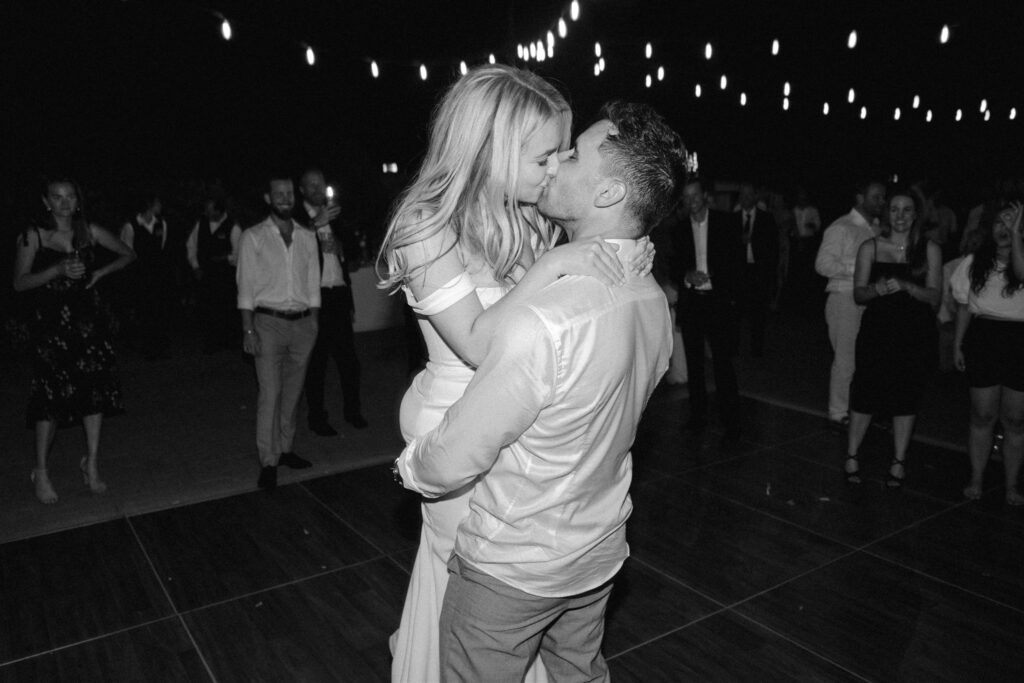 A couple kissing on a dance floor at night, surrounded by other people. String lights are hanging above, creating a festive atmosphere.