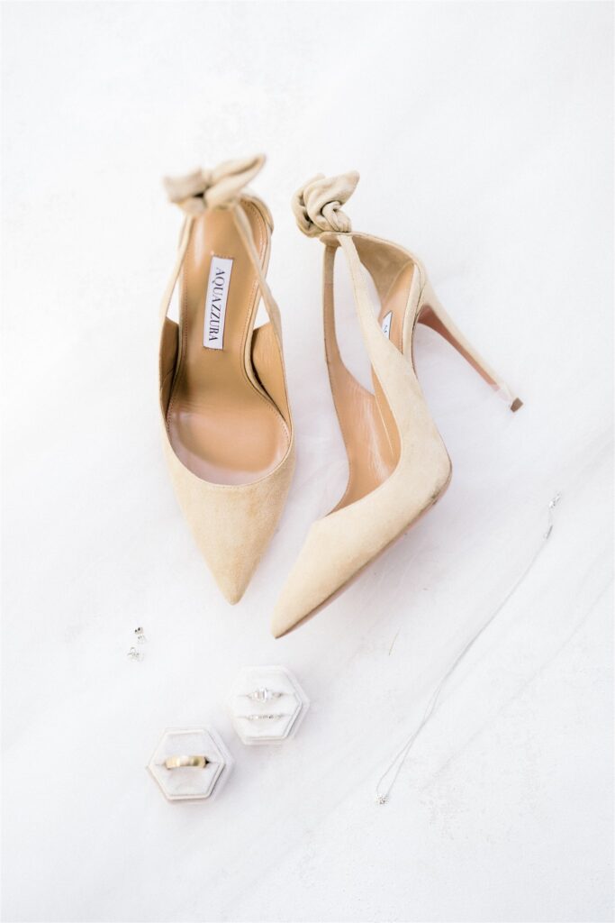 Beige high heels with bow accents next to two ring boxes containing rings and a thin chain bracelet on a white background.