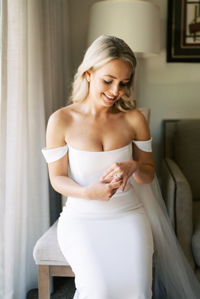 A woman in an off-the-shoulder white dress sits on a chair, looking down with a smile, holding a ring. A nearby lamp softly illuminates the scene.