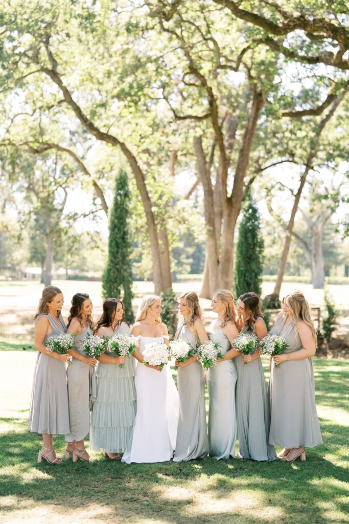 A bride in a white dress stands with eight bridesmaids in light gray dresses, holding bouquets, in a sunny outdoor setting with trees in the background.