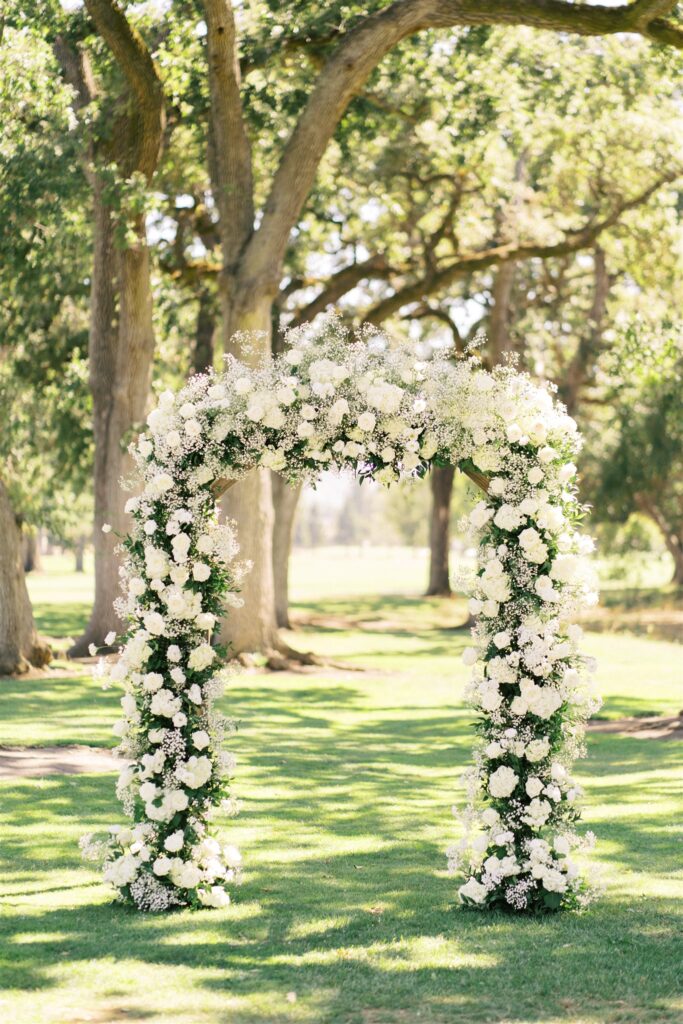 A floral arch made of white flowers stands on a grassy area under tall trees in a sunlit park.