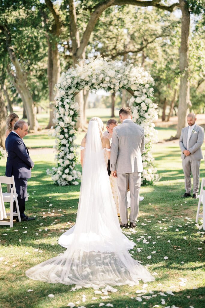 Bride and groom stand at an outdoor altar in front of a floral arch, surrounded by guests seated on white chairs, with scattered white petals on the grass.