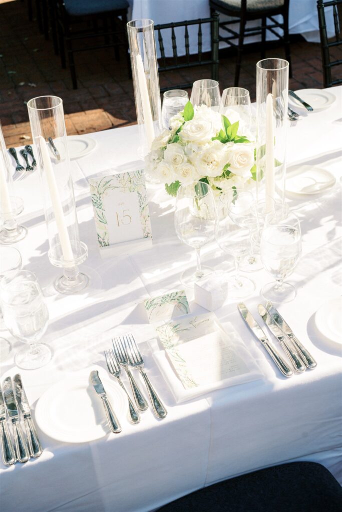 Elegant table setting with white roses, tall candles, glassware, and silver cutlery on a white tablecloth. Place card marked with the number 15.