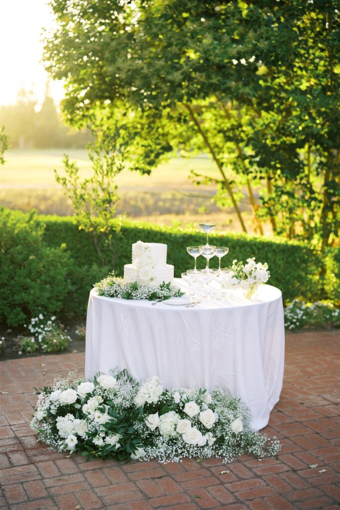 A white table with a tiered cake and stacked champagne glasses, surrounded by white flowers and greenery, is set on a brick patio with a green garden in the background.