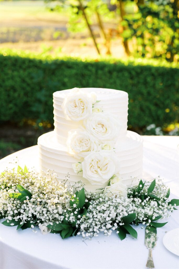 Two-tier white cake with ridged frosting, decorated with large white roses. Surrounded by baby's breath and greenery on a white tablecloth.