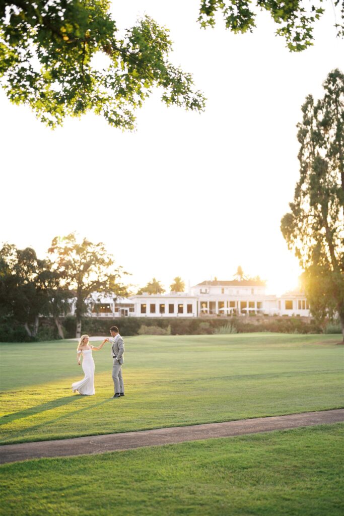 A couple dances on a grassy field at sunset, with a large building in the background.
