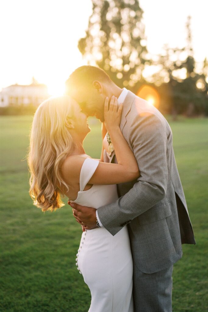 A couple embraces outdoors at sunset. The woman wears a white dress, and the man is in a gray suit. They are smiling with their foreheads touching, surrounded by a sunlit landscape.
