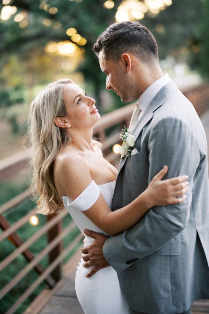A couple embraces and looks into each other's eyes on a wooden bridge, with trees and soft evening light in the background.