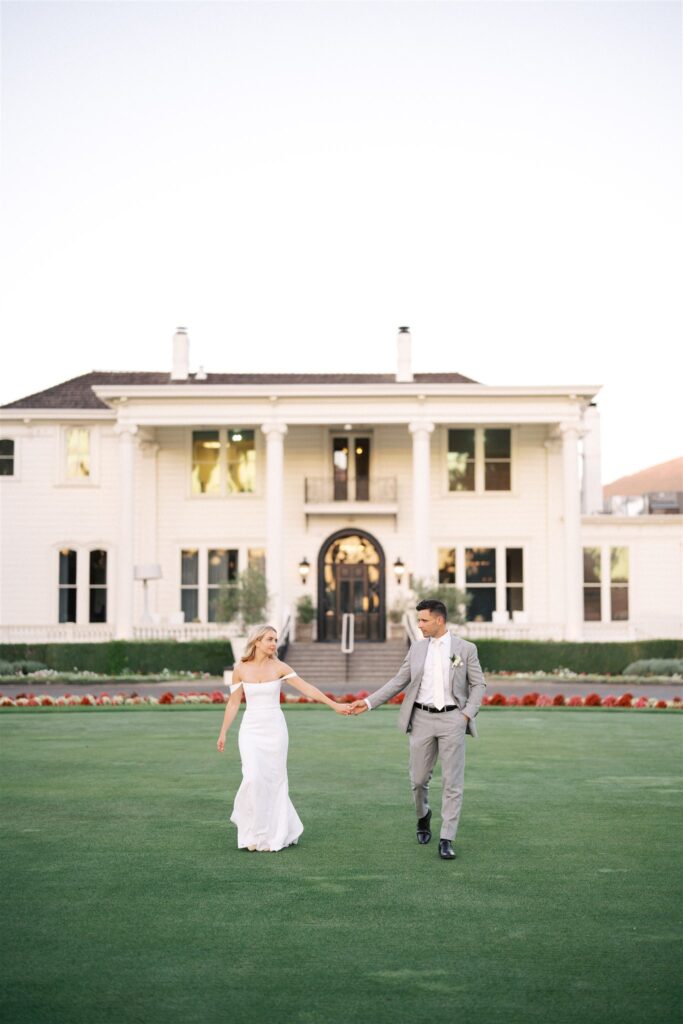 A couple walks hand-in-hand on a lawn in front of a large, white mansion with columns. The woman wears a white dress and the man wears a gray suit.