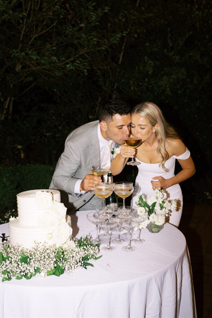 A couple in formal attire toast with champagne beside a tiered white cake on a decorated table, outdoors.