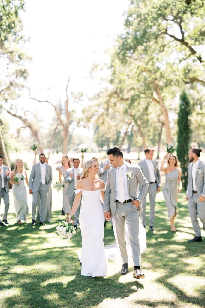 A bride and groom walk hand in hand on grass, surrounded by bridesmaids and groomsmen in gray attire, holding bouquets against a backdrop of trees.