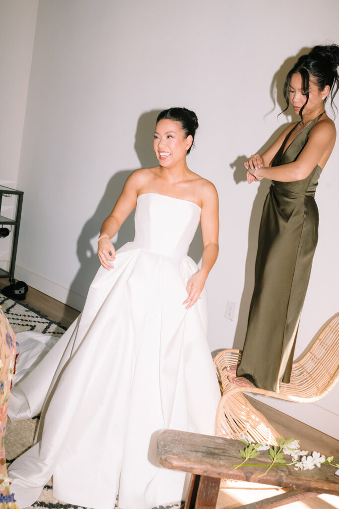 A woman in a strapless white wedding dress smiles while another woman in a green satin dress stands nearby adjusting her bracelet.