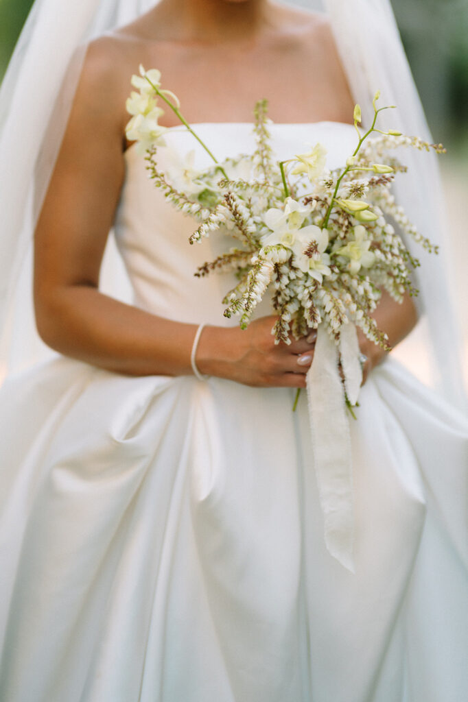 A bride in a strapless white gown holds a bouquet of white flowers and greenery, with a long white ribbon.