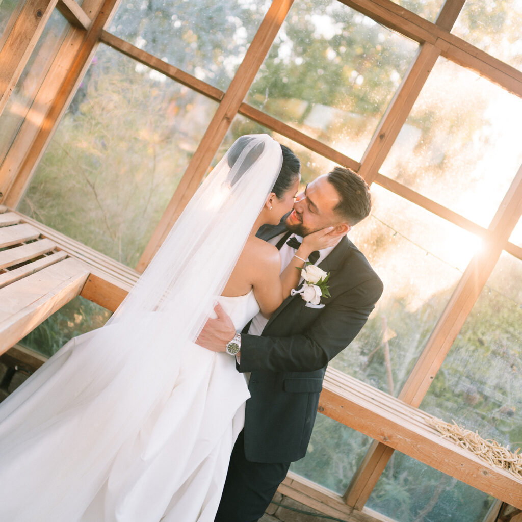 A bride and groom embrace and smile at each other in a sunlit wooden structure, with the bride wearing a long white veil and the groom in a black suit.