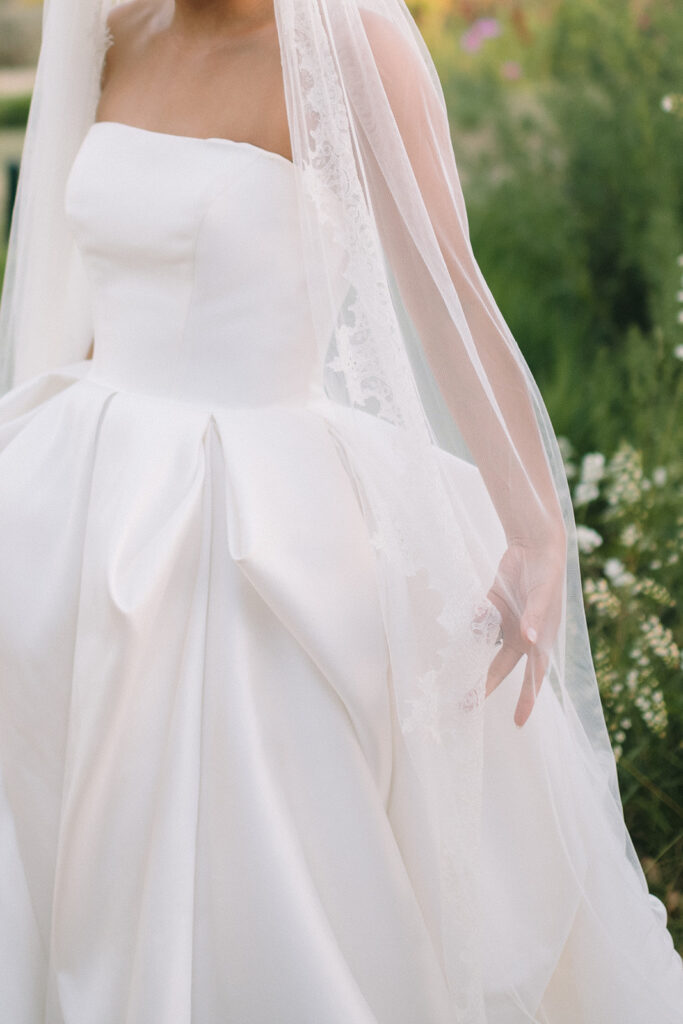 A bride in a strapless white wedding dress and lace-trimmed veil stands outdoors, surrounded by greenery and flowers.