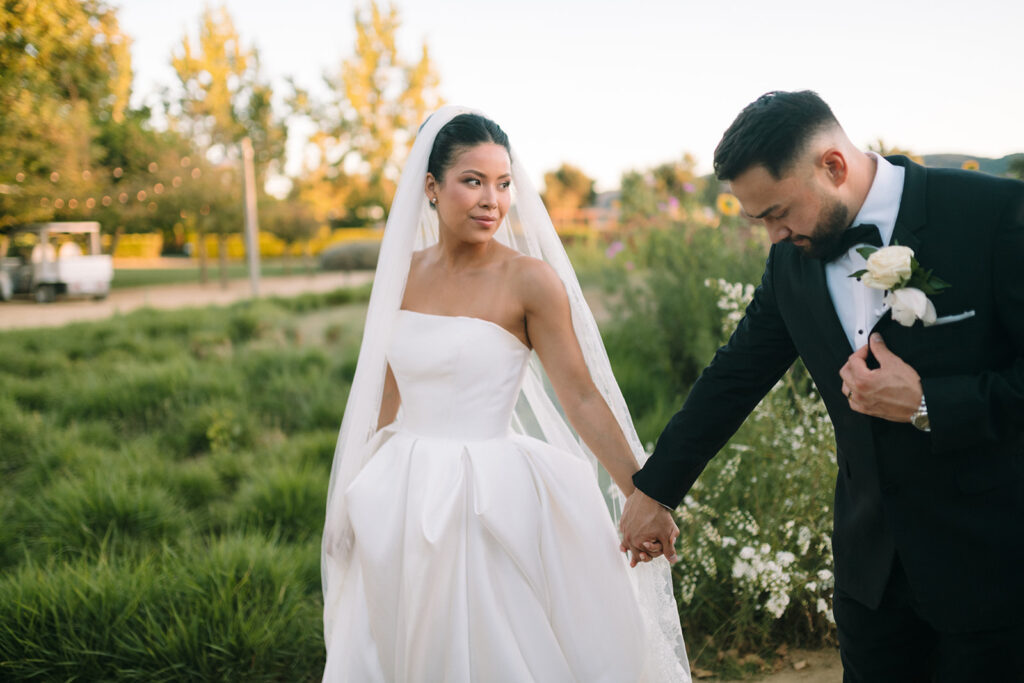 A bride in a white gown and a groom in a black suit hold hands outdoors, surrounded by greenery and flowers.