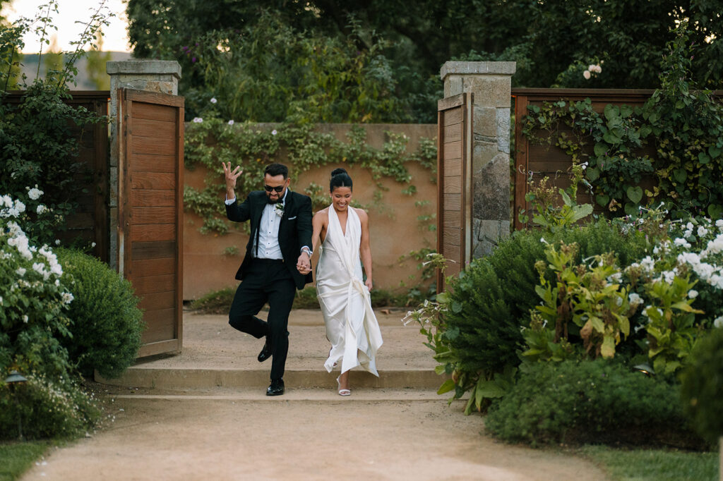 A couple in formal attire walks hand in hand down stone steps between open wooden gates, surrounded by lush greenery and flowers.