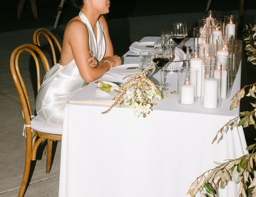 A woman in a white dress sits alone at a table set with candles, glassware, and a flower bouquet on a white tablecloth.