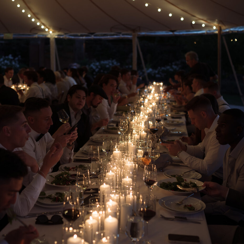 A long table under a tent is set for dinner with many people seated, eating and conversing, illuminated by numerous lit candles and string lights above.