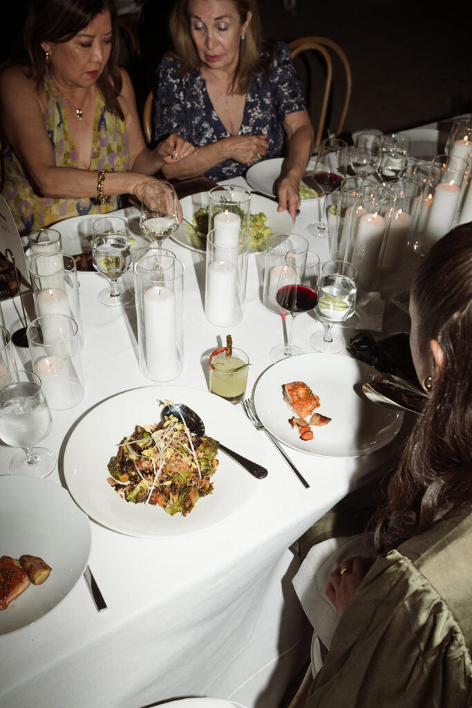 Three women sit at a candlelit dinner table set with salads, wine, cocktails, and water, engaged in conversation and serving food.