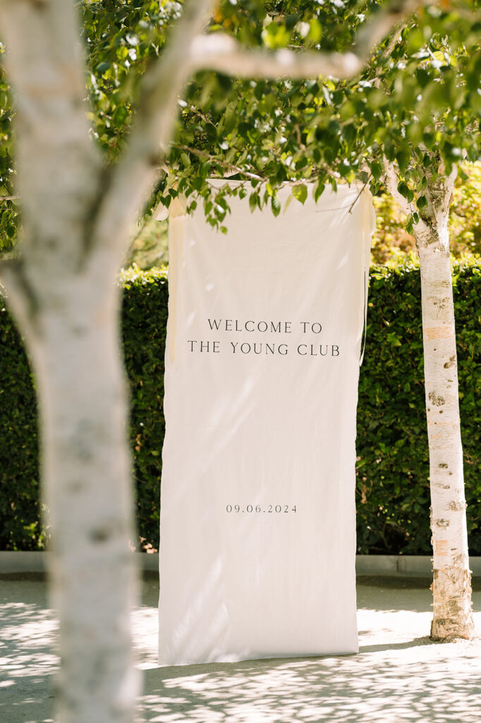 A cream-colored banner reading "WELCOME TO THE YOUNG CLUB" with the date "09.06.2024" is displayed outdoors between two trees.