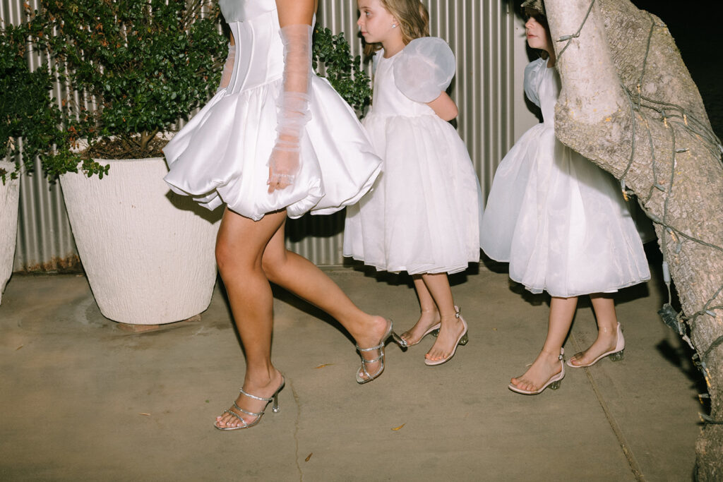Three people in white dresses and heeled sandals walk outside on a concrete path, with two young girls following an adult. Potted plants and a tree are visible in the background.