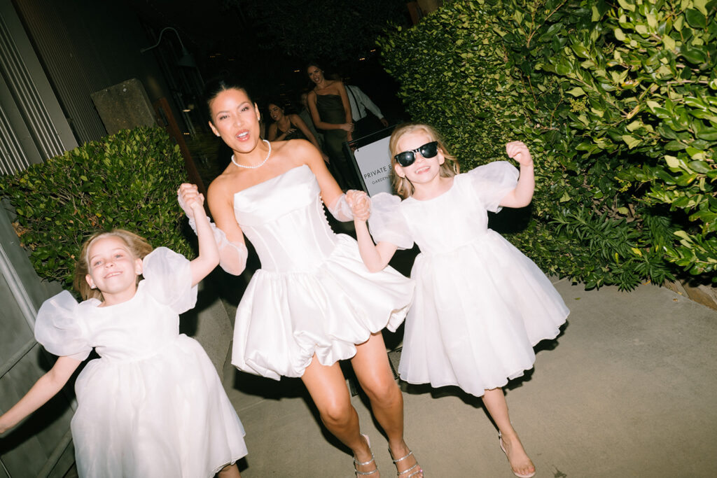 Three people wearing white dresses, including one adult and two children, walk together outdoors on a paved path lined with greenery at night.