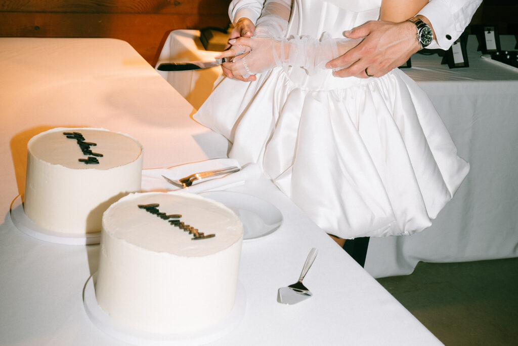 A couple, dressed in white, holds a knife together to cut one of two white frosted cakes on a table.