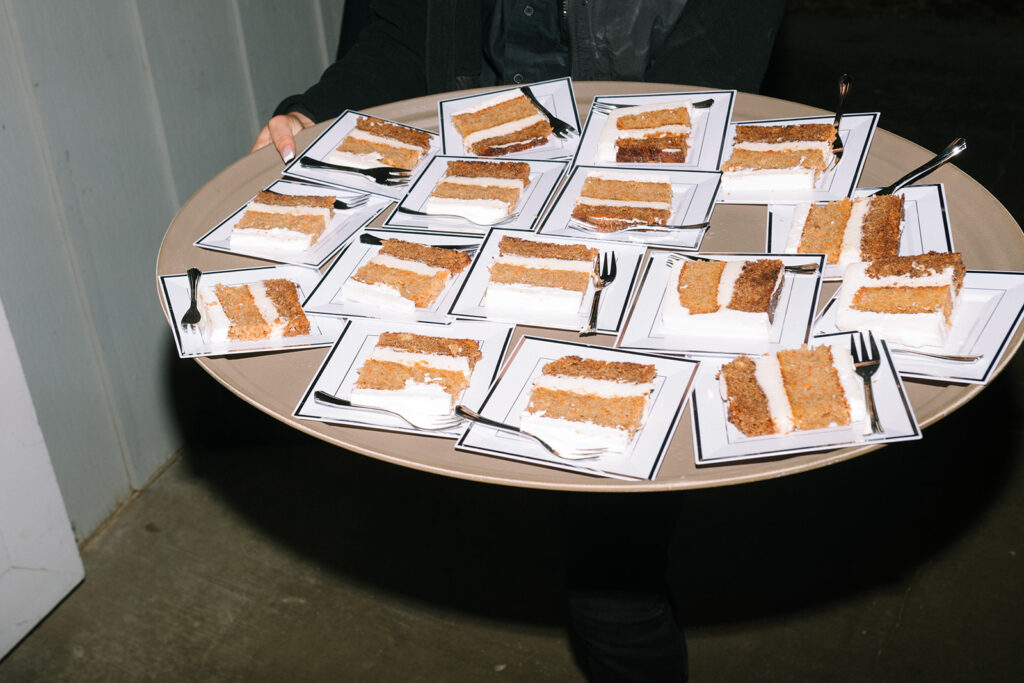 A person holds a large round tray with many square plates, each containing a slice of layered cake and a fork.