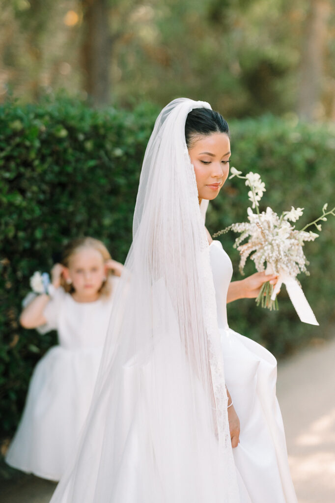 A bride in a white dress and veil holds a bouquet, while a young girl in a white dress stands in the background near green hedges.