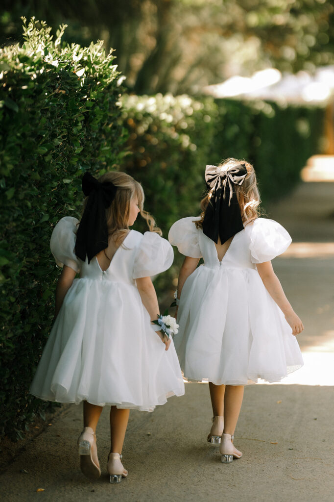 Two young girls in white dresses with puffed sleeves and large black hair bows walk side by side on a sunny path next to a green hedge.