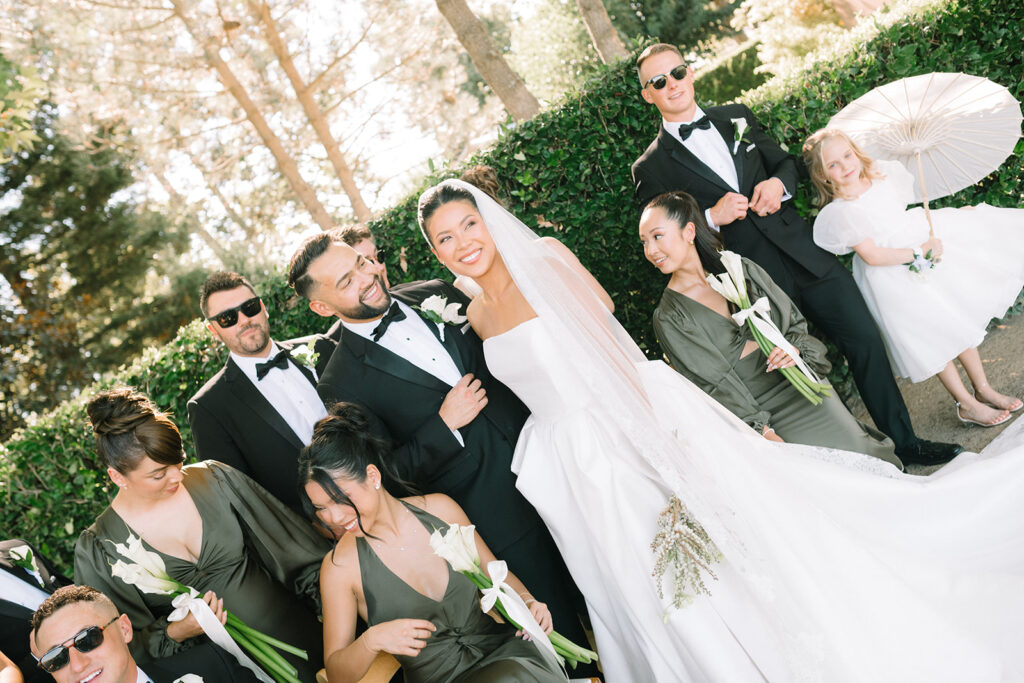 A bride and groom stand outdoors with their wedding party dressed in black and green, holding white flowers; a young girl with a parasol stands to the side.
