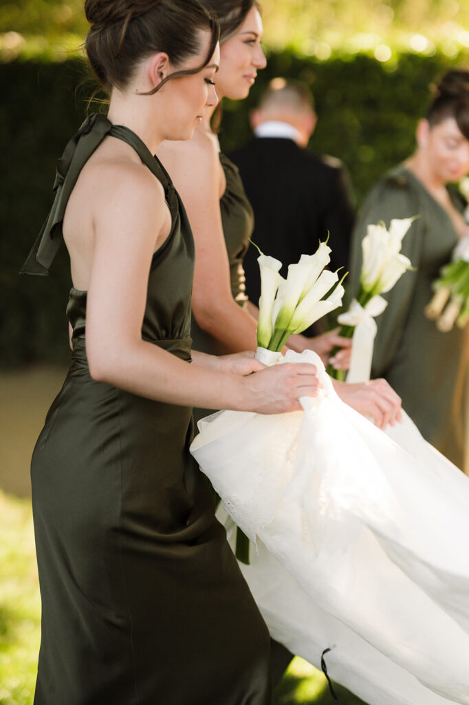 Two women in dark green dresses hold white calla lilies and assist with a white wedding dress outdoors, while another person stands in the background.