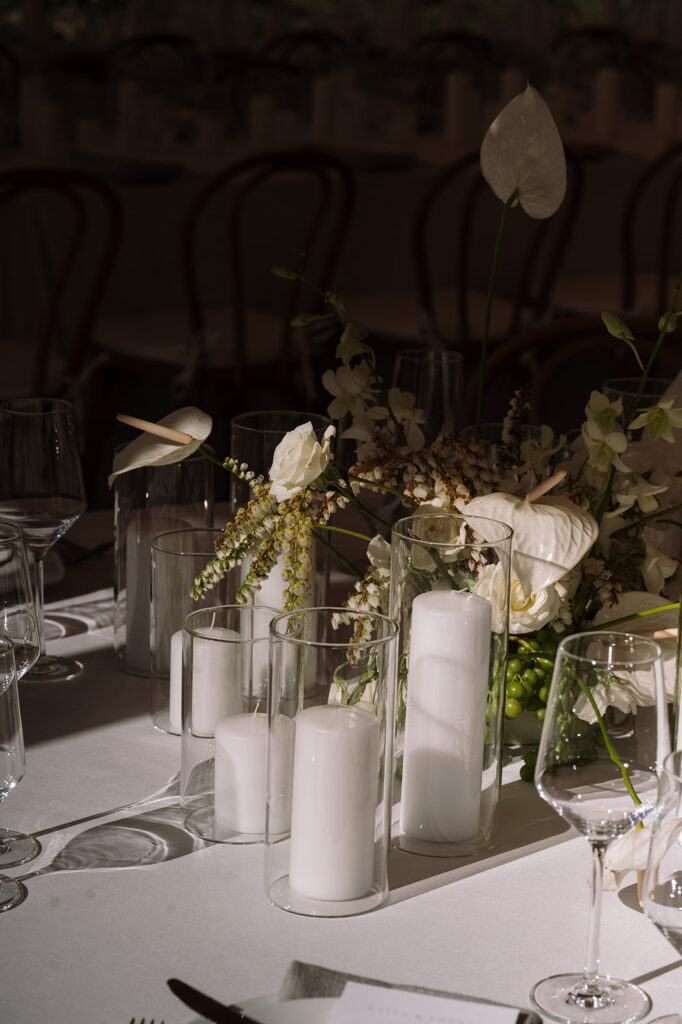 A close-up of a table set with white pillar candles in glass holders, white flowers, and glassware on a white tablecloth.
