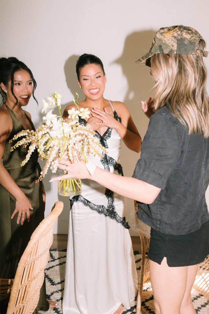 Three women indoors; one in a white dress with black trim holding flowers, another in a green dress, and a third in casual clothes with a camouflage hat.