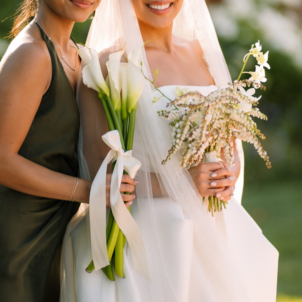 Two women pose together outdoors; one in a white wedding dress and veil holding a bouquet, the other in a dark dress holding calla lilies tied with ribbon.