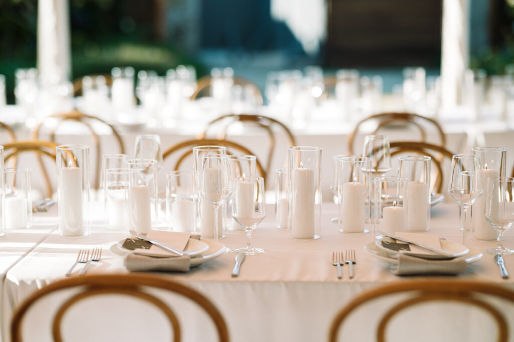 A formal table setting with white tablecloths, multiple clear glasses, white plates, napkins, cutlery, and white pillar candles, arranged for an event or reception.